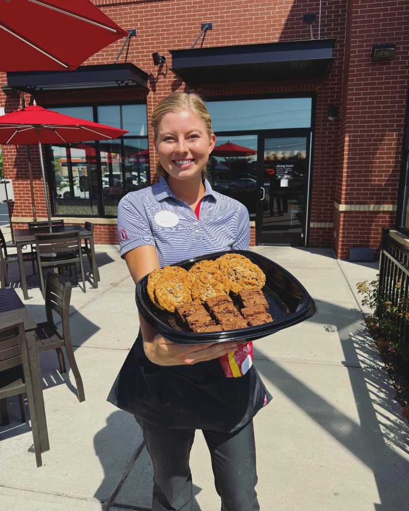 Smiling Chick-fil-A team member holding a Chocolate Chunk Cookie and Chocolate Fudge Brownie tray outside a restaurant
