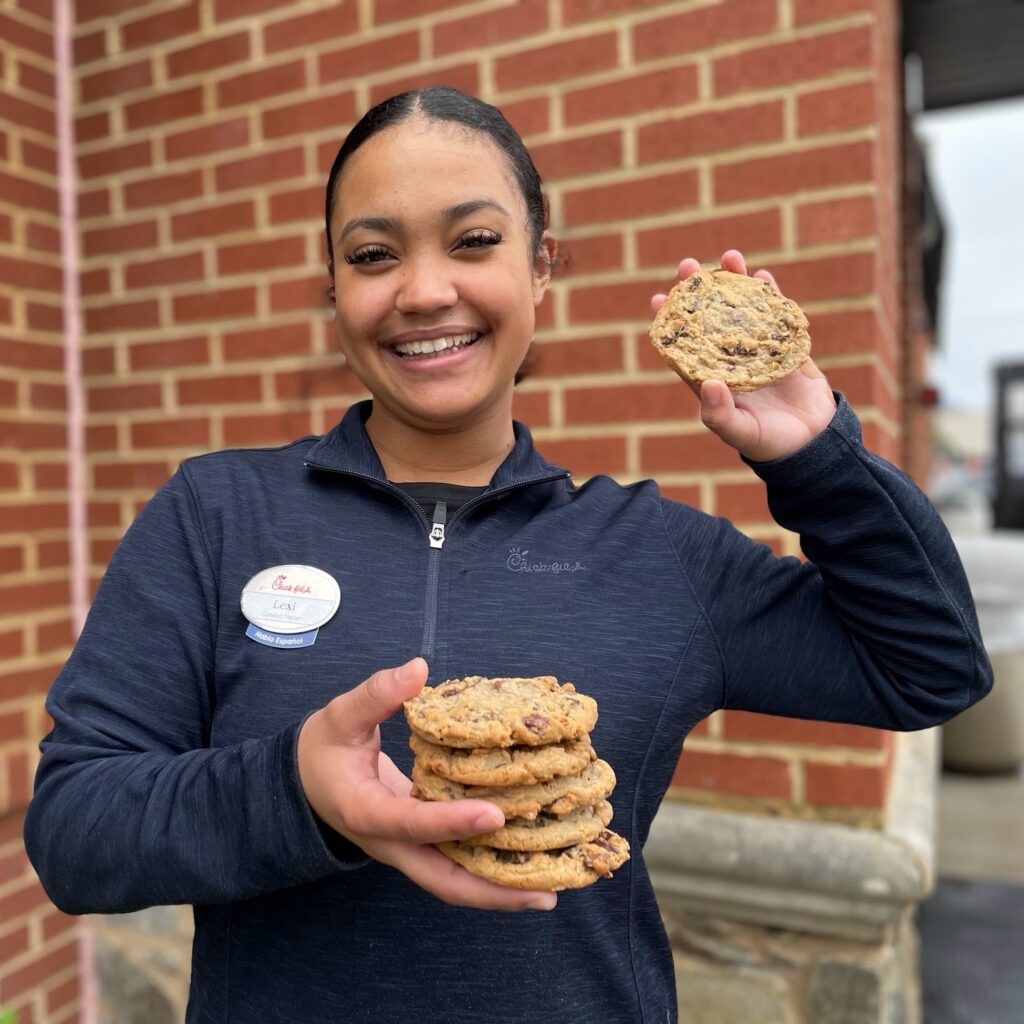 Smiling Chick-fil-A team member holding up a Chocolate Chunk Cookie and a stack of cookies