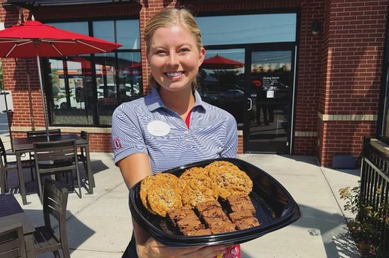 Smiling Chick-fil-A team member holding a Chocolate Chunk Cookie and Chocolate Fudge Brownie tray outside a restaurant
